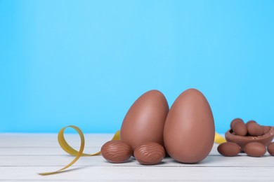 Delicious chocolate eggs and golden ribbon on white wooden table against light blue background