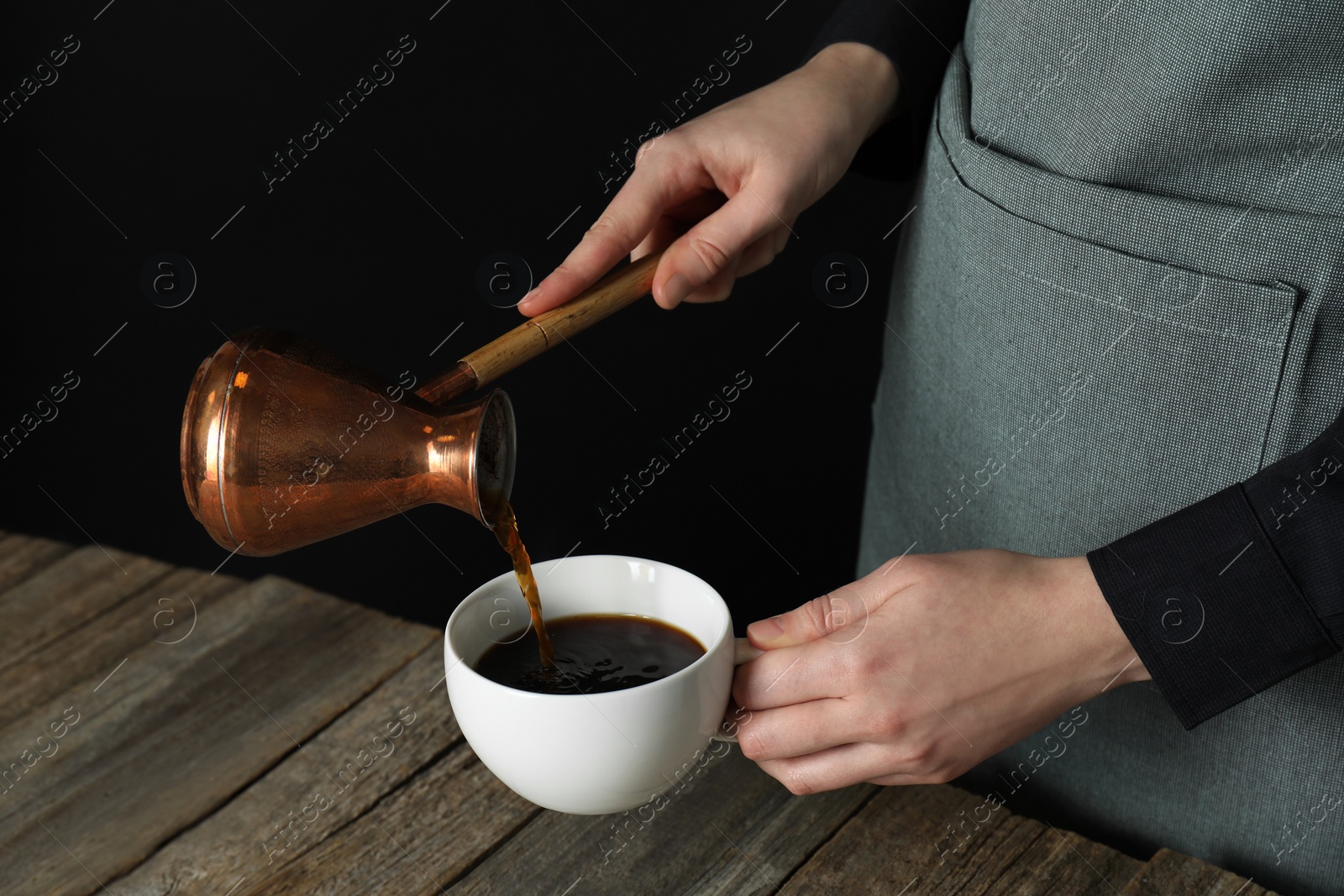 Photo of Turkish coffee. Woman pouring brewed beverage from cezve into cup at wooden table against black background, closeup