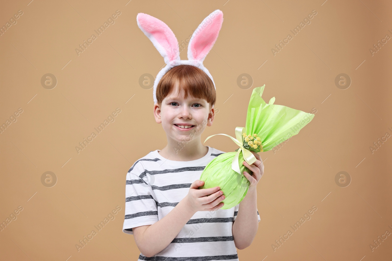 Photo of Easter celebration. Cute little boy with bunny ears and wrapped egg on dark beige background