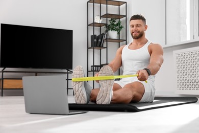 Photo of Muscular man doing exercise with elastic resistance band near laptop on mat at home