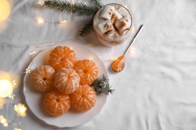 Peeled delicious ripe tangerines, festive lights and glass of drink with marshmallows on white bedsheet, flat lay. Space for text