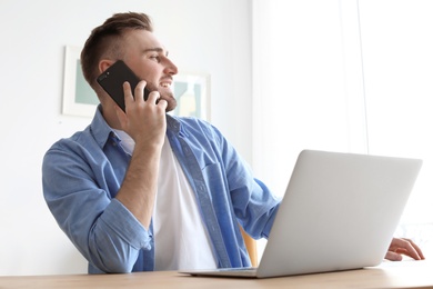 Photo of Young man talking on mobile phone while working with laptop at desk. Home office