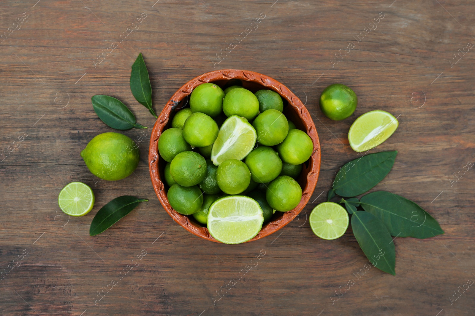 Photo of Whole and cut fresh ripe limes in bowl on wooden table, flat lay