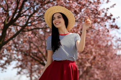 Photo of Pretty young woman with straw hat near beautiful blossoming trees outdoors. Stylish spring look