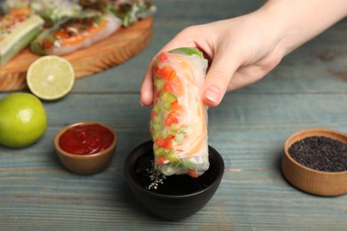 Woman dipping delicious roll wrapped in rice paper into soy sauce at blue wooden table, closeup