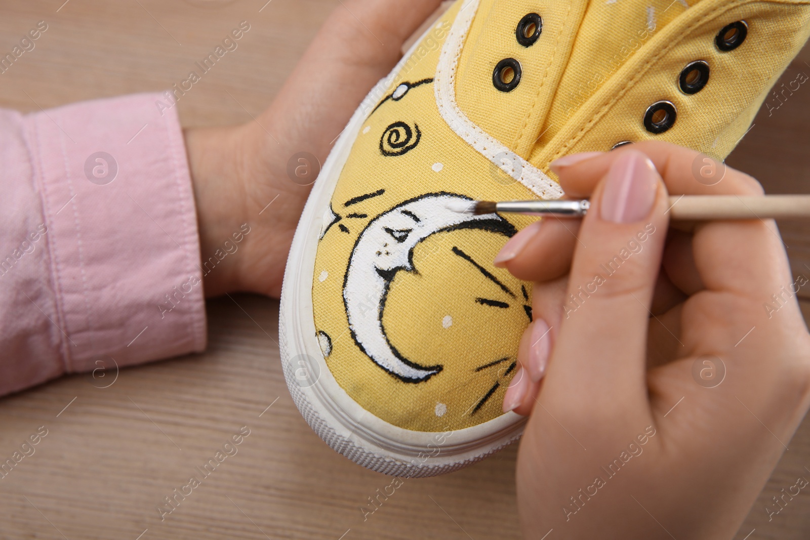 Photo of Woman painting on sneaker at wooden table, closeup. Customized shoes