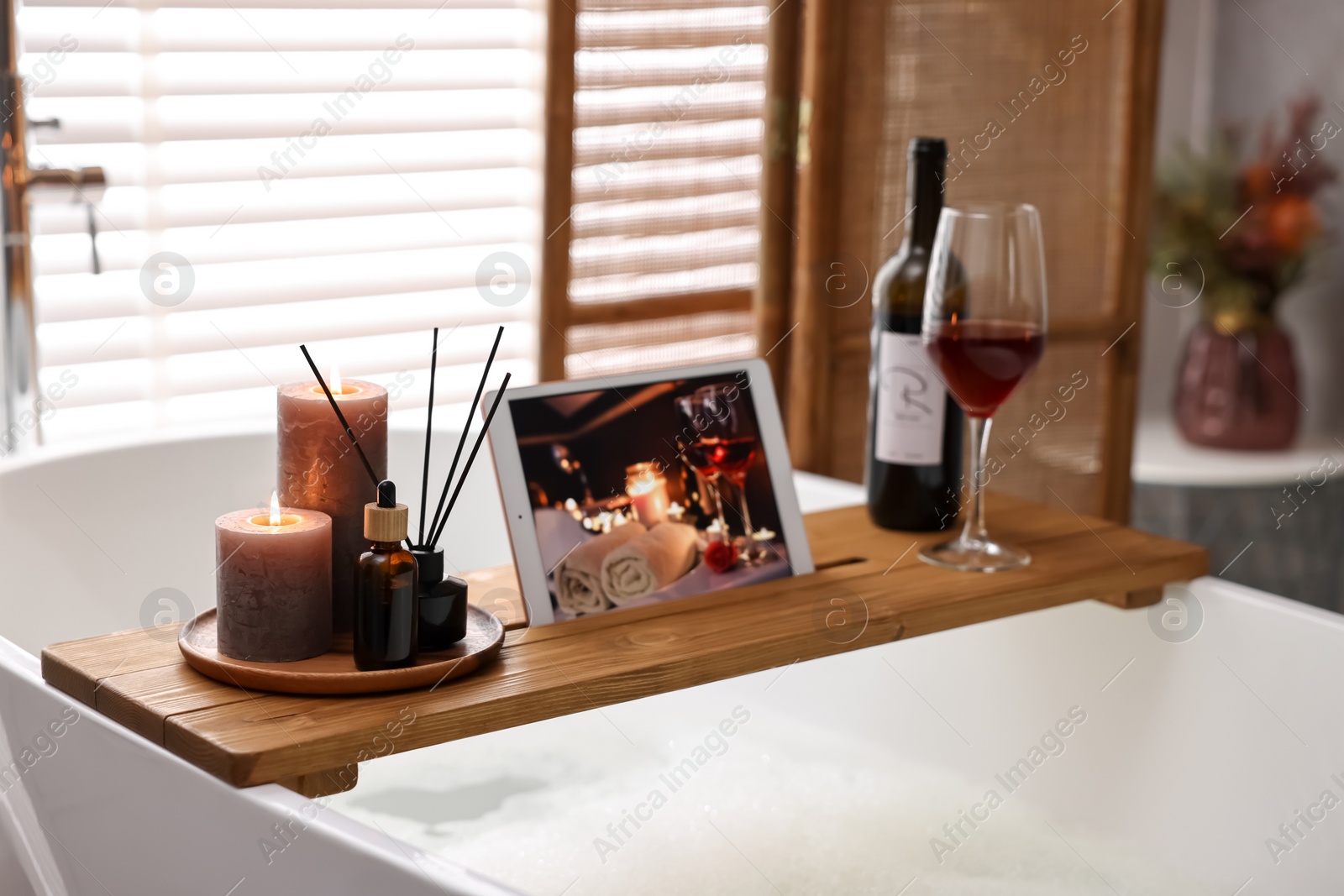 Photo of Wooden tray with tablet, wine and burning candles on bathtub in bathroom