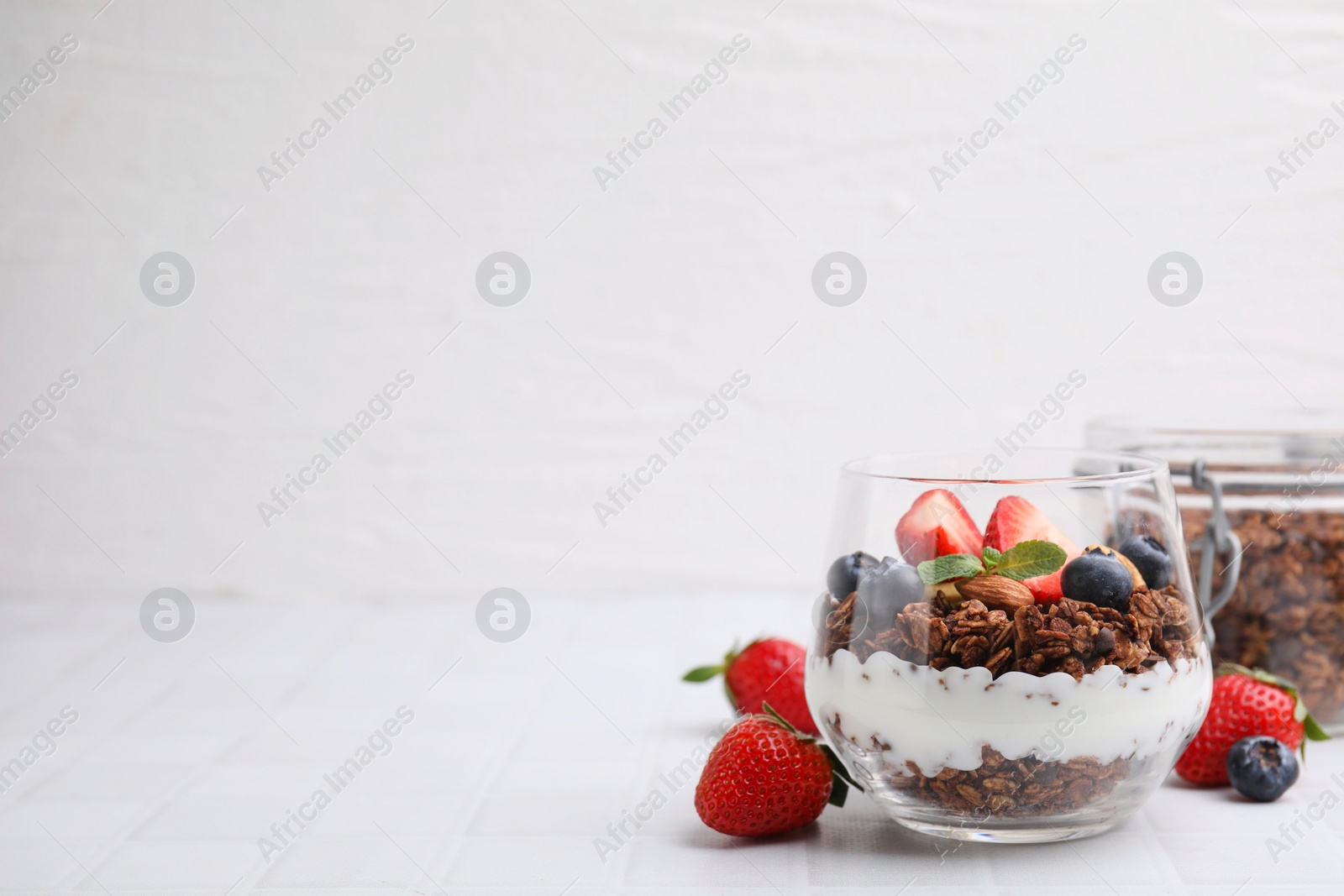 Photo of Tasty granola with berries, yogurt and mint in glass on white tiled table, space for text