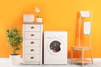 Beautiful bathroom interior with washing machine, stylish furniture and houseplants near bright orange wall