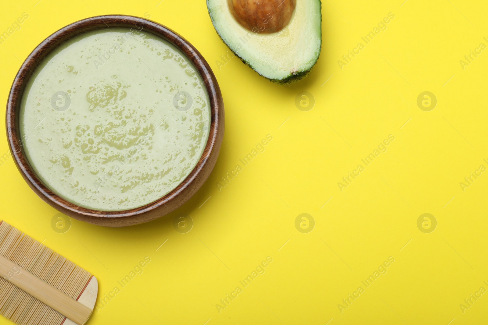 Photo of Homemade hair mask in bowl, avocado and bamboo comb on yellow background, flat lay. Space for text