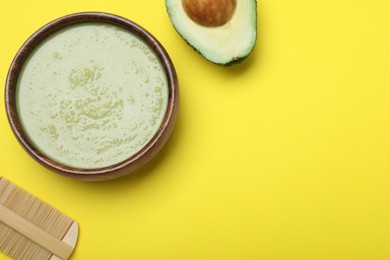 Homemade hair mask in bowl, avocado and bamboo comb on yellow background, flat lay. Space for text