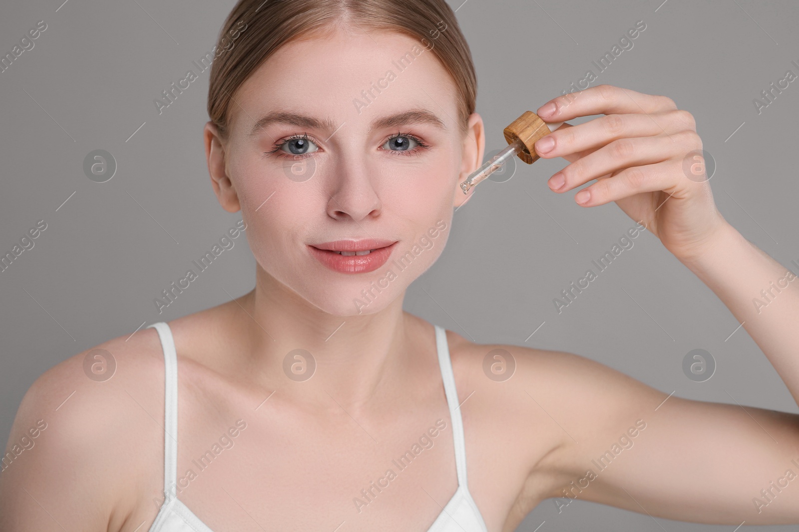 Photo of Beautiful young woman applying essential oil onto face on grey background