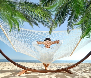 Image of Man relaxing in hammock under green palm leaves on sunlit beach