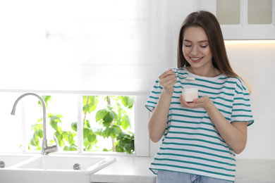 Young attractive woman eating tasty yogurt in kitchen. Space for text