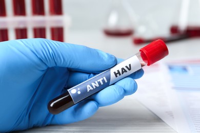 Scientist holding tube with blood sample and label Anti HAV at white wooden table, closeup