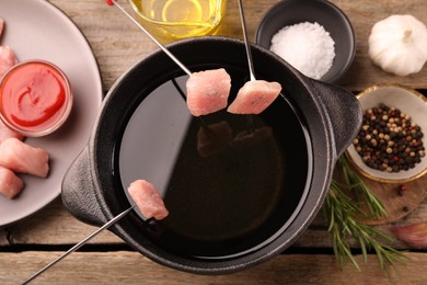 Fondue pot with oil, forks, raw meat pieces and other products on wooden table, flat lay