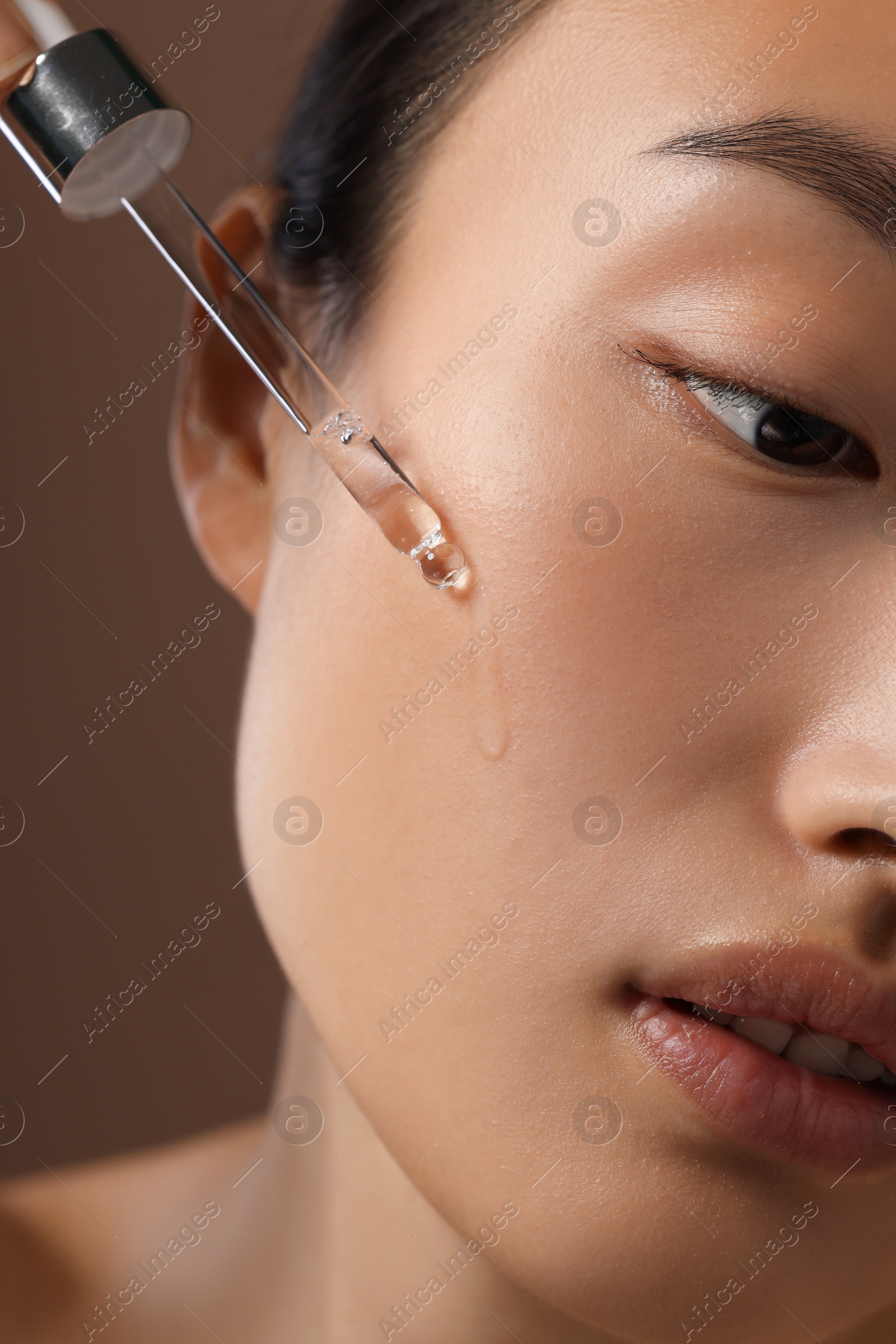 Photo of Beautiful young woman applying cosmetic serum onto her face on brown background, closeup