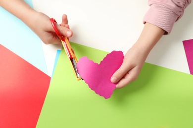 Photo of Child cutting out paper heart with craft scissors at table, top view