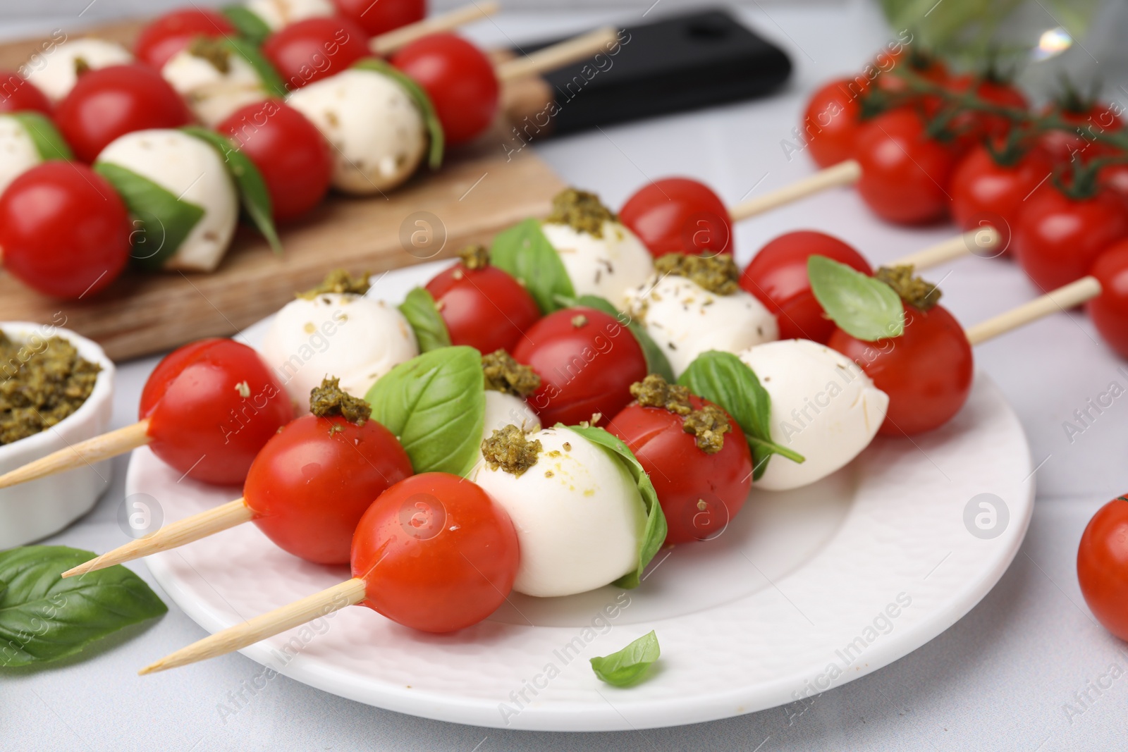 Photo of Caprese skewers with tomatoes, mozzarella balls, basil and pesto sauce on white table, closeup