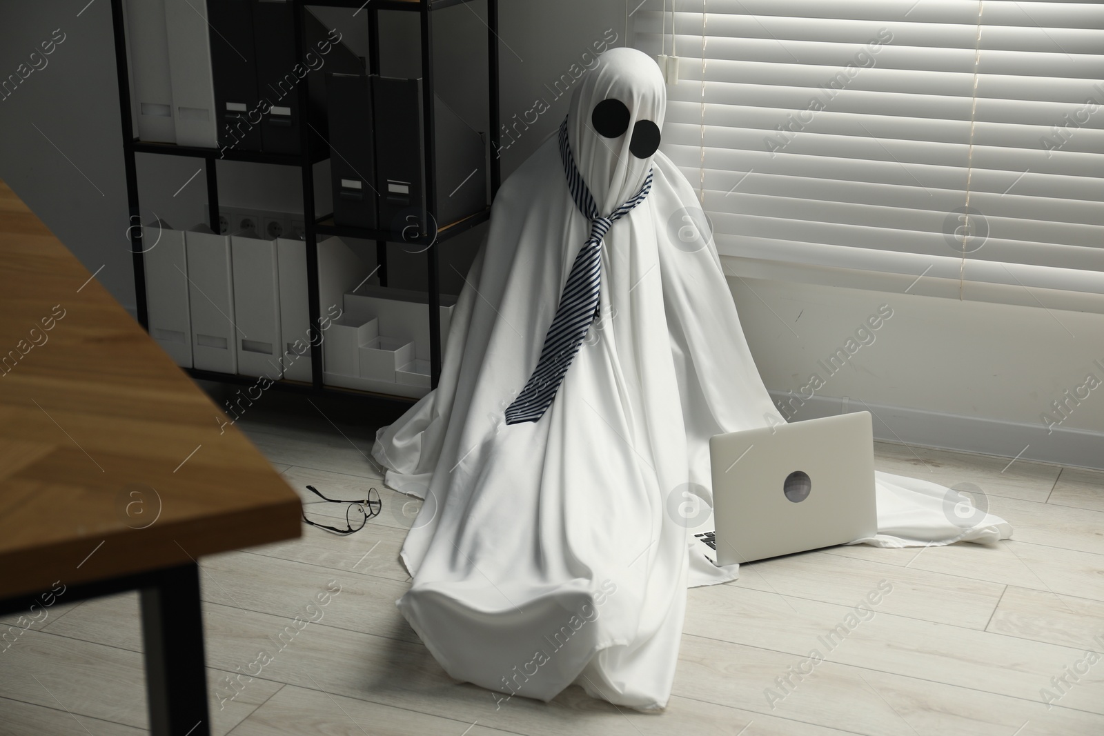 Photo of Overworked ghost. Man in white sheet with laptop on floor in office