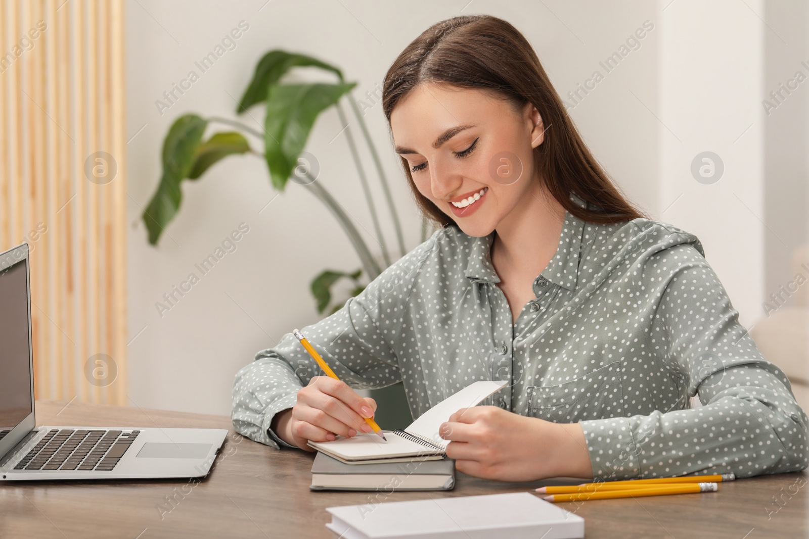 Photo of Happy young woman with notebook working on laptop at wooden table indoors