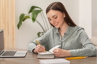 Happy young woman with notebook working on laptop at wooden table indoors