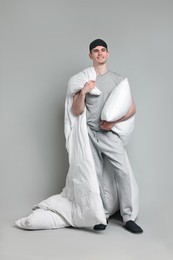 Happy man in pyjama holding pillow and blanket on grey background