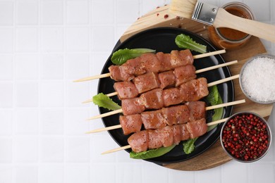 Photo of Wooden skewers with cut raw marinated meat , spices and basting brush on white tiled table, flat lay. Space for text