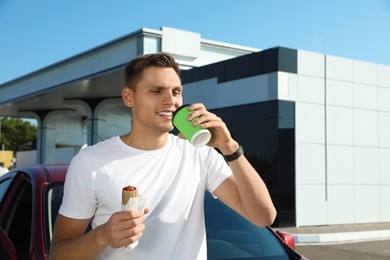 Photo of Young man with hot dog drinking coffee near car at gas station