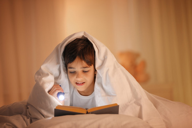Boy with flashlight reading book under blanket at home