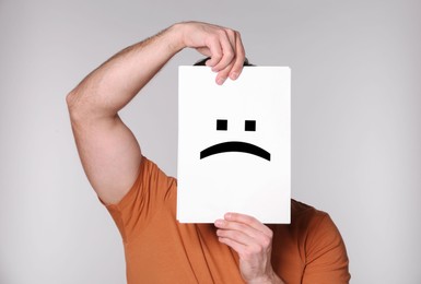 Man hiding behind sheet of paper with sad face on grey background