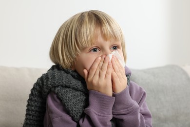 Photo of Boy blowing nose in tissue on sofa in room. Cold symptoms