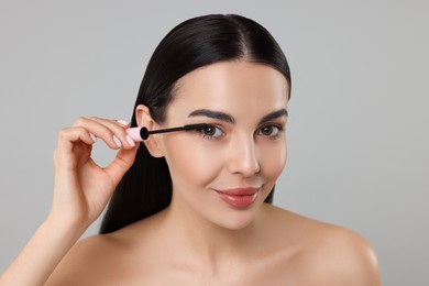 Beautiful young woman applying mascara on grey background