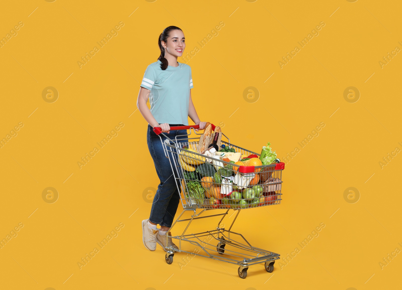 Photo of Happy woman with shopping cart full of groceries on yellow background