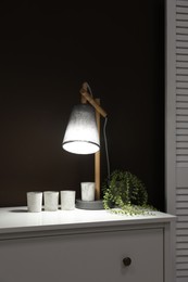 Stylish lamp, decor and green plant on white chest of drawers near brown wall indoors. Interior design