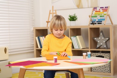 Photo of Cute little boy cutting orange paper at desk in room. Home workplace