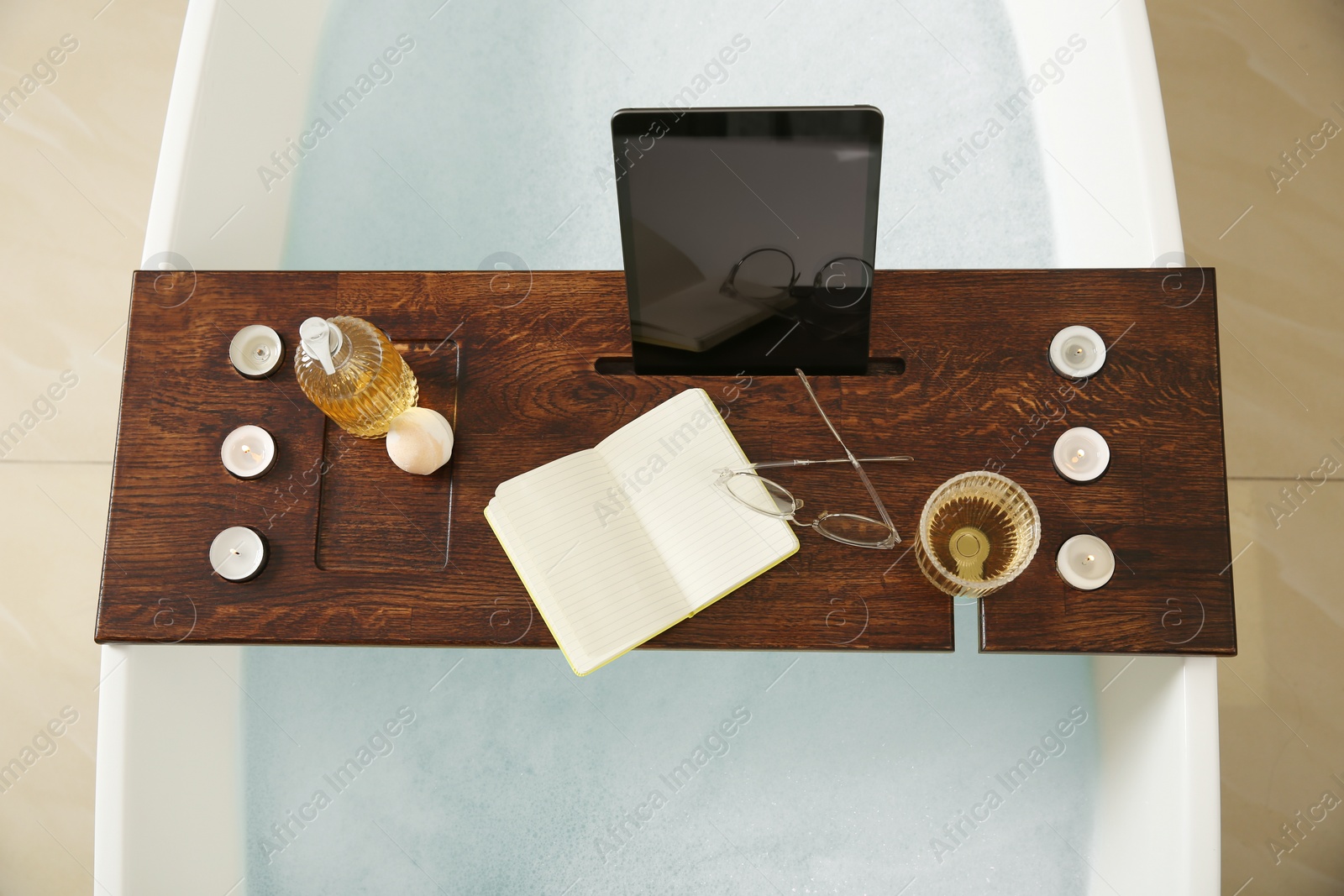 Photo of Wooden bath tray with candles, book and tablet on tub indoors, top view