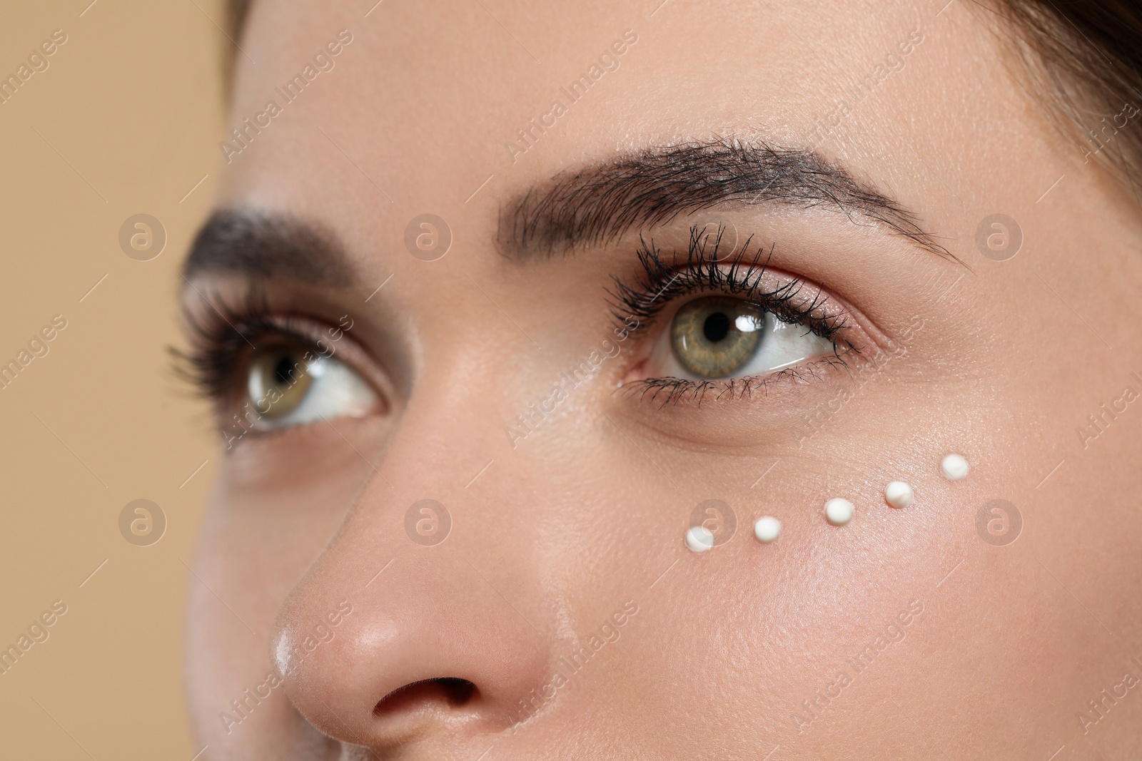 Photo of Young woman with cream around eye on beige background, closeup