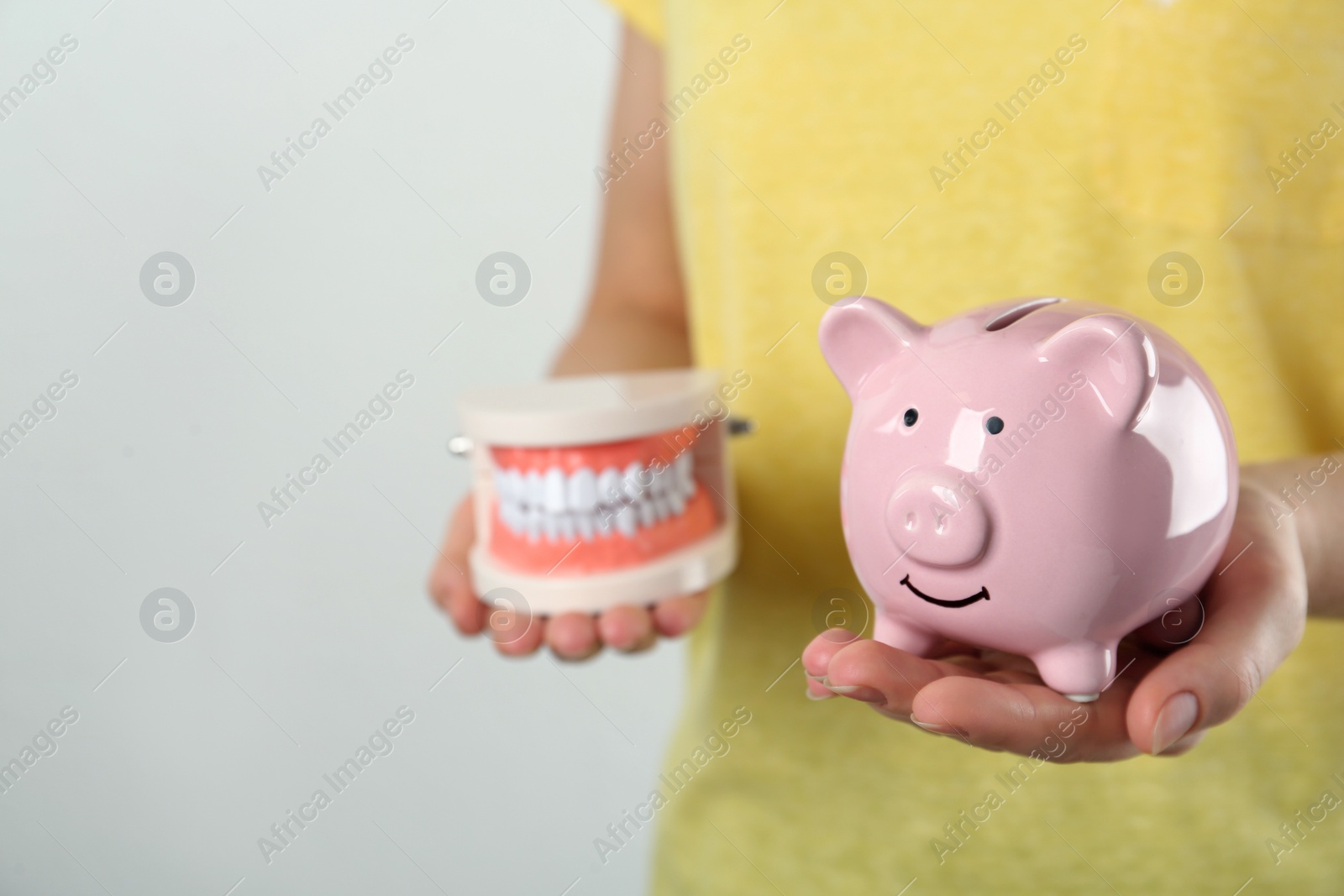 Photo of Woman holding educational dental typodont model and piggy bank on light background, closeup. Expensive treatment