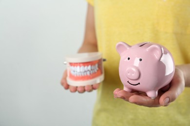 Woman holding educational dental typodont model and piggy bank on light background, closeup. Expensive treatment