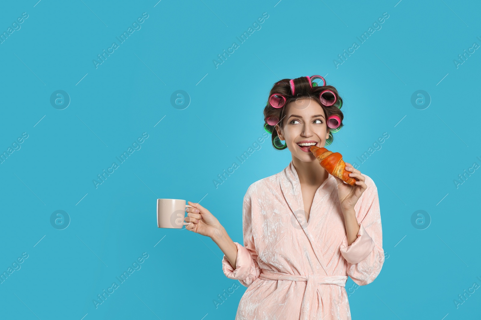 Photo of Happy young woman in bathrobe with hair curlers holding croissant and cup of drink on light blue background. Space for text