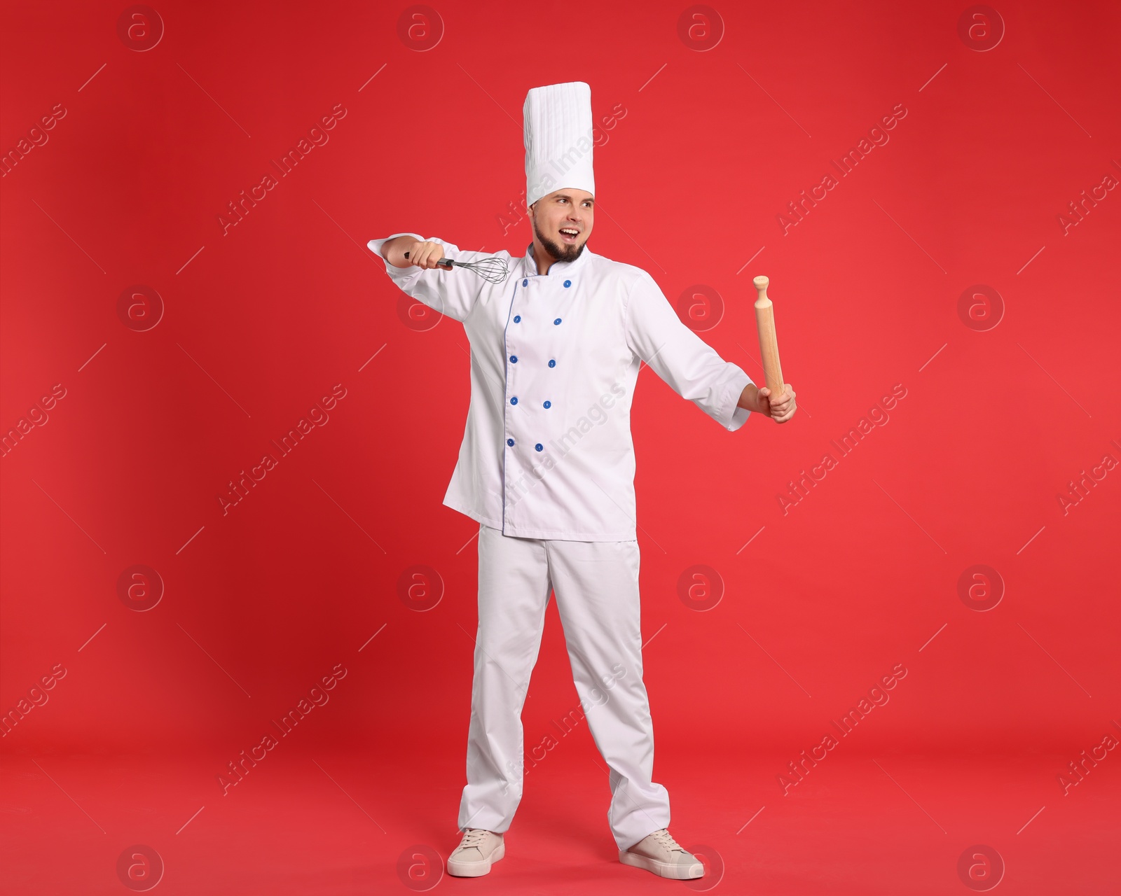 Photo of Happy professional confectioner in uniform holding rolling pin and whisk on red background