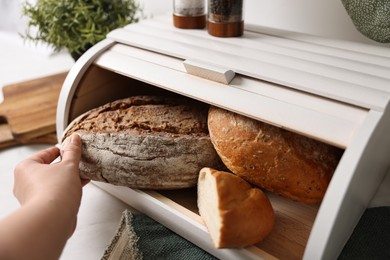 Photo of Woman taking loaf from wooden bread basket at white table in kitchen, closeup