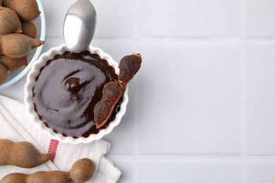 Photo of Bowl of tamarind sauce, spoon and fresh pods on white tiled table, flat lay. Space for text