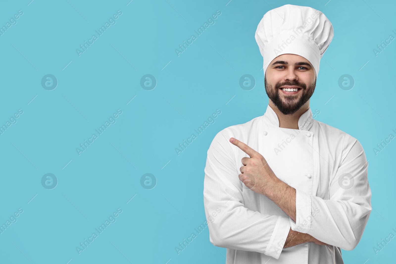 Photo of Happy young chef in uniform pointing at something on light blue background. Space for text