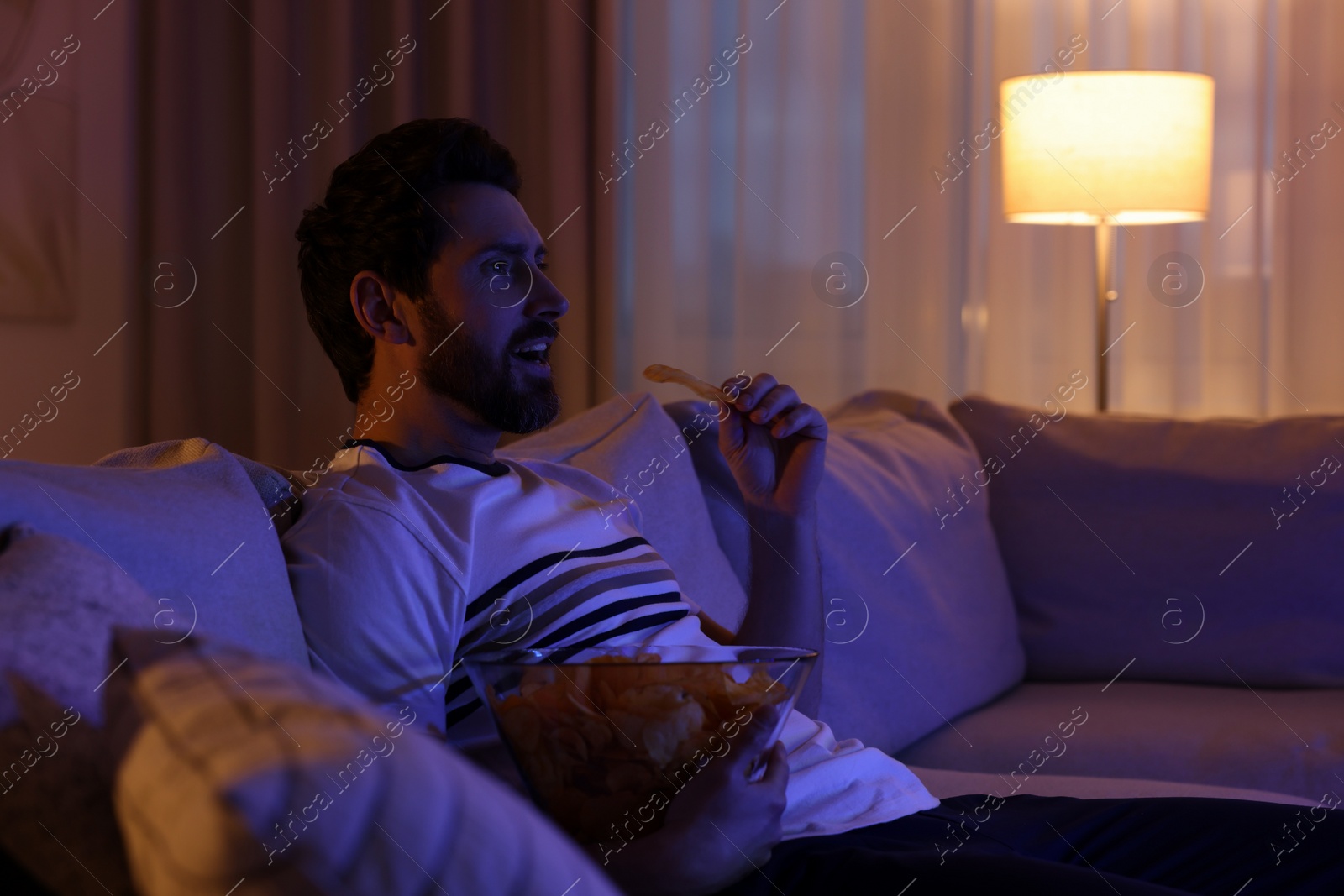 Photo of Man eating chips while watching TV on sofa at night. Bad habit