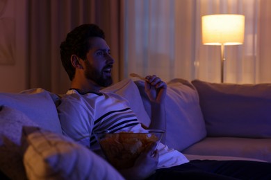 Photo of Man eating chips while watching TV on sofa at night. Bad habit