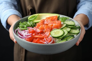 Woman holding delicious poke bowl with salmon and vegetables on black background, closeup