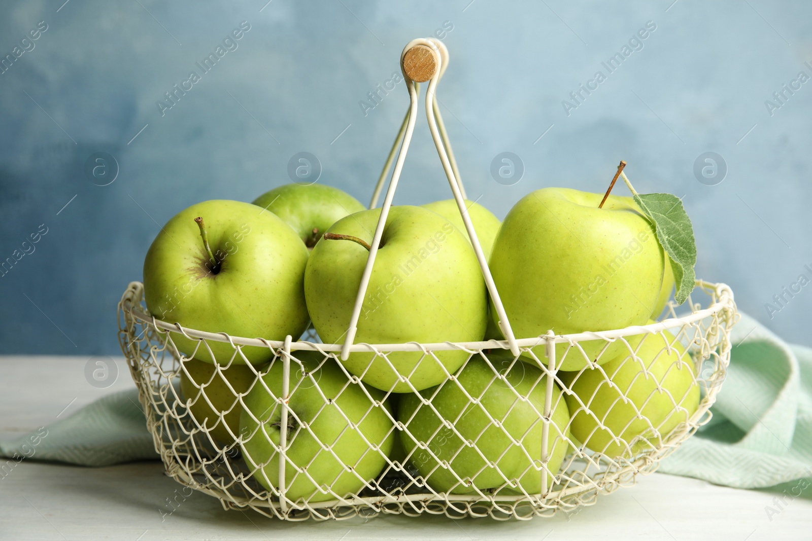 Photo of Metal basket of fresh ripe green apples on white wooden table against blue background