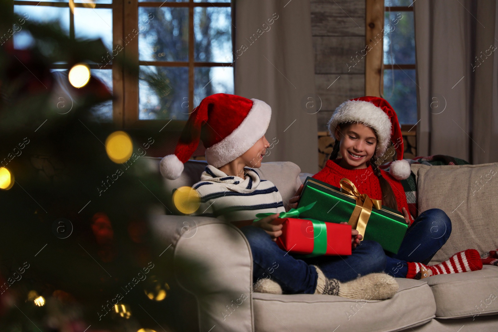Photo of Happy children wearing Santa hats with Christmas gifts on sofa at home